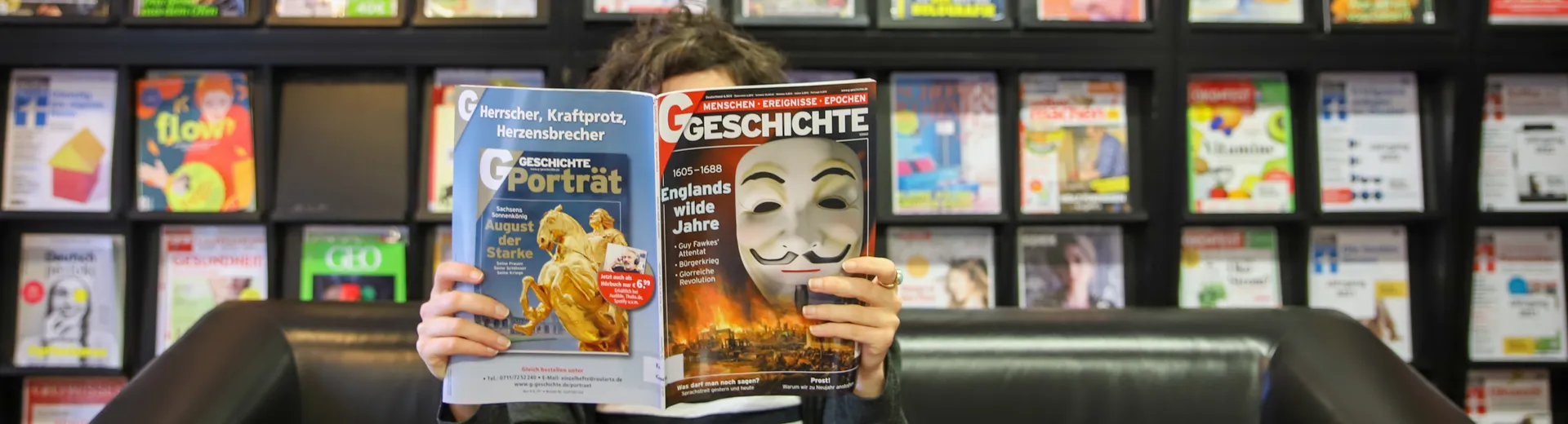Lesende Person vor einem Bücherregal, das Gesicht ist von einer aufgeschlagenen Zeitschrift verdeckt. Verteilseite Stadtbücherei