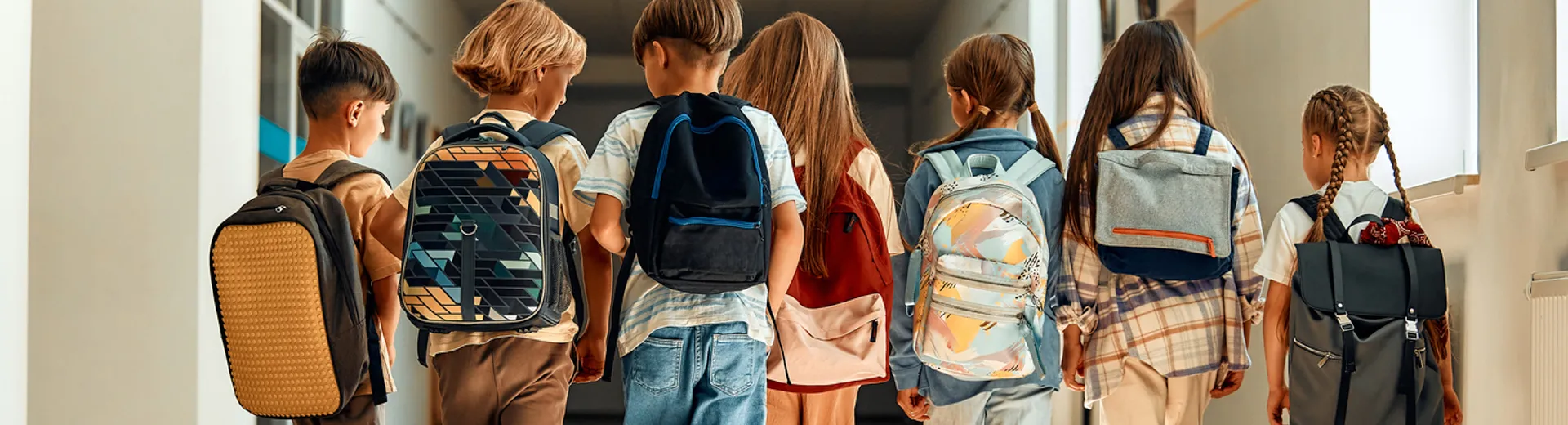 Gruppe von Kindern mit Ranzen und Rücksäcken läuft durch einen Schulflur, von hinten fotografiert. Schule und Bildung