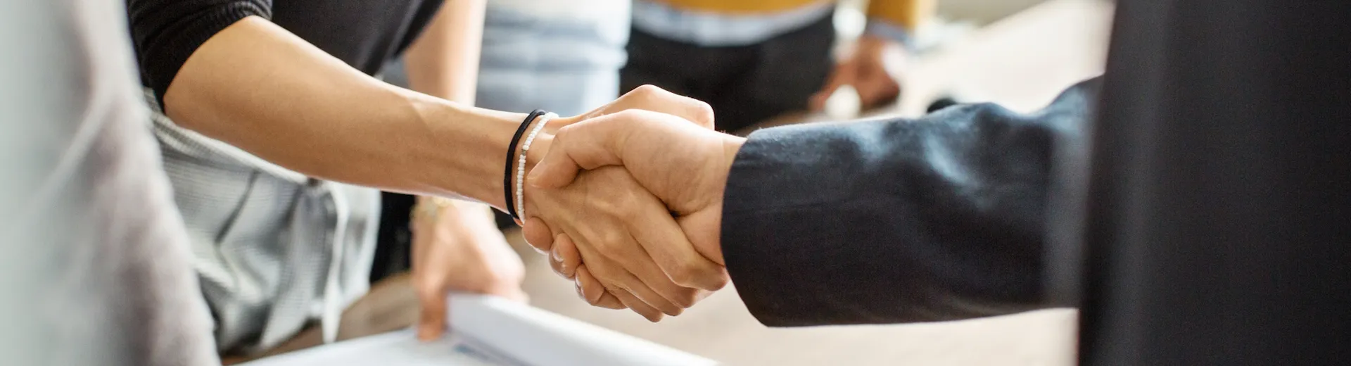 Nahaufnahme von Menschen, die an einem Tisch stehen. Zwei Personen schütteln sich die Hände. Verteilseite Wirtschaftsförderung shake hands