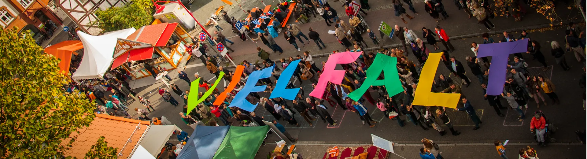 Luftaufnahme: In der Schulstraße findet das Fest zum 3. Oktober statt. Besucher*innen zeigen mit großen bunten Buchstaben das Wort Verteilseite Integration, Schulstraße, 3. Oktober, Vielfalt