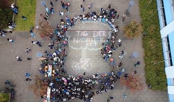 Auf dem Schulhof wurde mit Straßenkreide ein Fahrrad mit einem Weg gemalt. Um das Bild sind viele Personen versammelt.