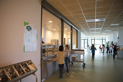 In der Cafeteria sind Kinder die ihr Mittagessen entgegennehmen und sich an die Esstische setzten.