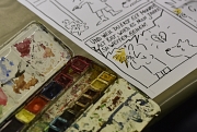 In einer Detailaufnahme ist ein Aquarell-Kasten zu sehen. Daneben werden diese Farben genutzt, um einen Comic zu kolorieren.