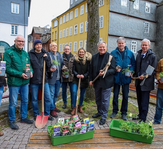 Eine Gruppe von Menschen steht im Halbkreis auf einer gepflasterten Fläche. einige haben Schippen, andere kleine Pflanzen in der Hand.