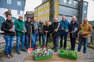 Eine Gruppe von Menschen steht im Halbkreis auf einer gepflasterten Fläche. einige haben Schippen, andere kleine Pflanzen in der Hand.