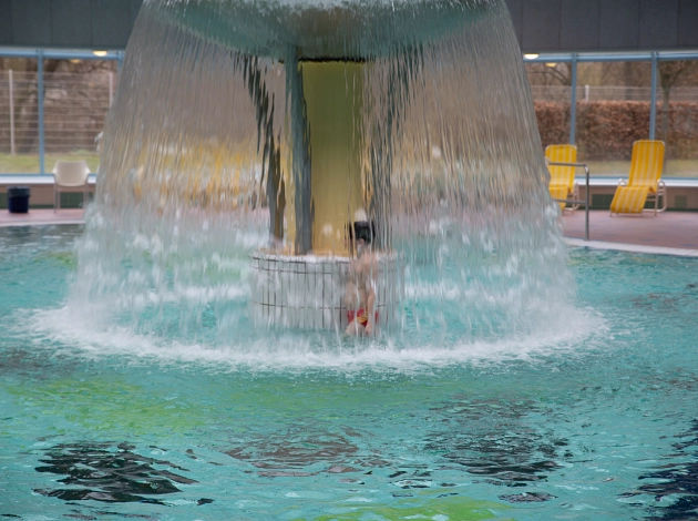 Kind in roter Badehose läuft unter dem Wasserpilz im Aktionsbecken des Marburger Sport- und Freizeitbades AquaMar durch..
