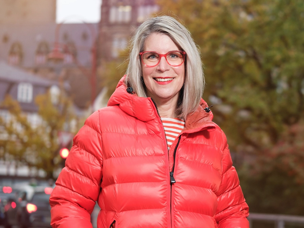 Porträt einer Frau mittleren Alters mit blonden halblangen Haaren und roter Brille.