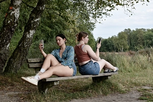 Ausstellung_Was heißt Einsamkeit für dich Zwei Frauen sitzen Rücken an Rücken auf einer Parkbank und halten jeweils ein Handy in der Hand.