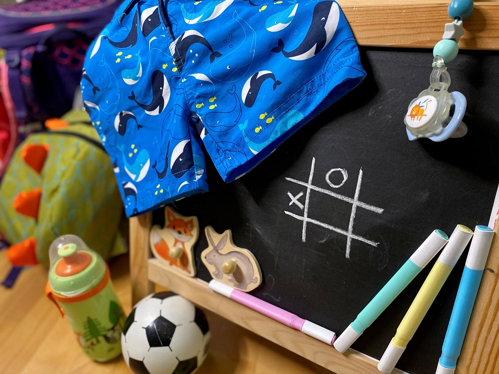 Betreuungsangebote für Kinder und Jugendliche Mehrere Gegenstände, die Kindern und Jugendlichen zugeordnet werden können: Eine Kindertafel, auf der mit Kreide ein Tic-Tac-Toe-Spiel aufgemalt ist eine Badeshorts, ein Schnuller an einer Schnullerkette, Filzstifte, Puzzleteile, ein Ball, eine Trinkflasche, ein kleines Boot, im Hintergrund ein Schulranzen und ein Kinder-Rucksack. © Simone Batz, Stadt Marburg