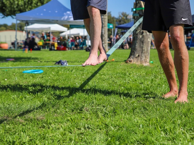 Eine Person balanciert auf einer Slackline, eine Person unterstützt. Zu sehen sind nur die Füße.