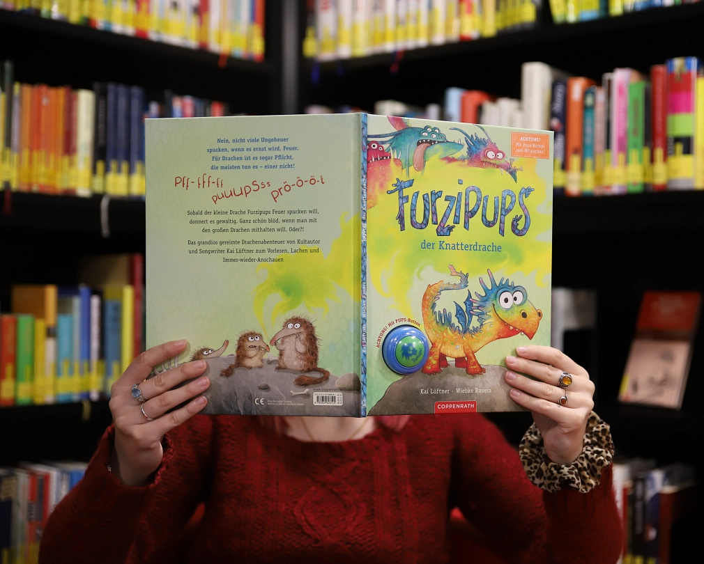 Zwei Hände halten das Buch „Furzipups, der Knatterdrache“ hoch. Die Person dahinter ist nicht zu sehen, nur ein Teil des Oberkörpers. Im Hintergrund stehen Bücherregale.&nbsp;&copy;&nbsp;Stefanie Ingwersen, Stadt Marburg