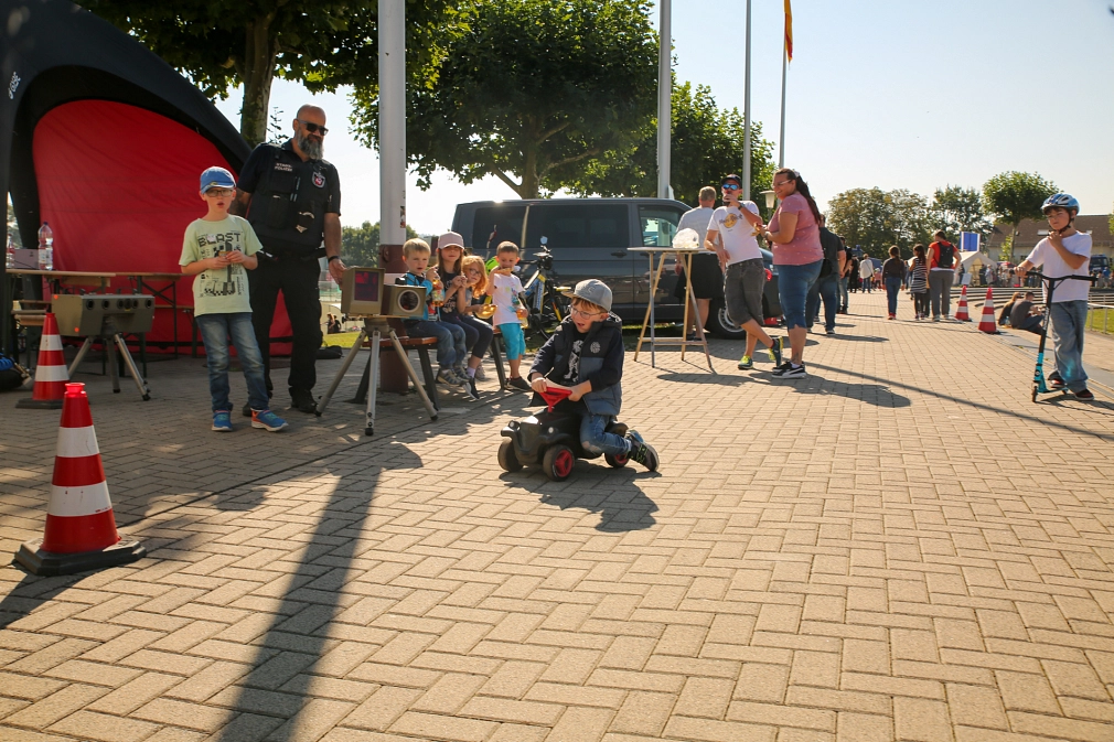 Das Bild zeigt ein Kind auf einem Bobbycar. Um ihn herum stehen noch weitere Menschen.