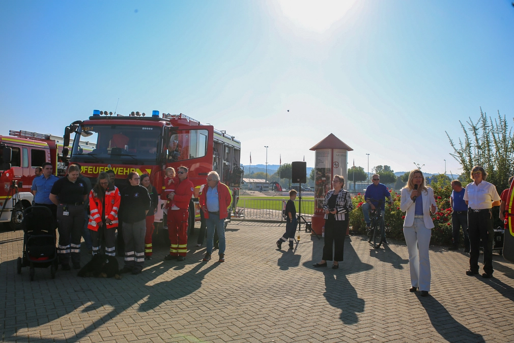 Das Bild zeigt links ein Feuerwehrauto, vor dem Menschen stehen, und rechts zwei Frauen, die ein Mikrofon in der Hand halten und hinein sprechen.
