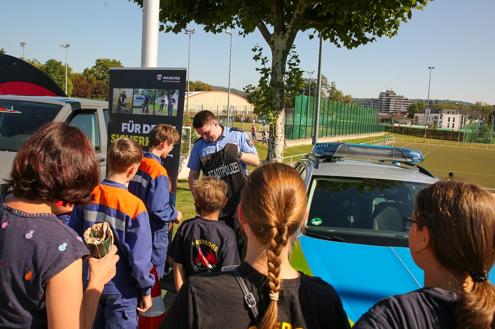 Das Bild zeigt mehrere Menschen vor einem Polizeiwagen, die einem Polizisten in Uniform zuhören.