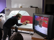 Ein Junge steht neben einem Tisch und bereitet die nächste Aufnahme für den animierten Lego-Trick-Film vor. Rechts steht ein Fernseher, in dem man das Geschehen ein zweites mal sehen kann.