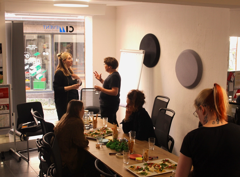 CIM_Creativity and Social Innovation Hub Marburg_Foto_Hannah Spitzer Innenansicht vom CIM: mehrere Personen im Gespräch. Zwei stehen am großen Fenster zur Straße, zwei sitzen am gedeckten Tisch. © Hannah Spitzer
