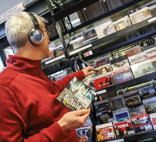 Das digitale Angebot der Stadtbücherei umfasst unter anderem Hörbücher Ein Mann steht in der Stadtbücherei vor einem Regal mit Hörbüchern und trägt Kopfhörer. Er hält eine CD in der Hand.