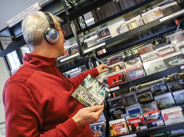 Ein Mann steht in der Stadtbücherei vor einem Regal mit Hörbüchern und trägt Kopfhörer. Er hält eine CD in der Hand.