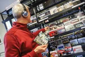 Das digitale Angebot der Stadtbücherei umfasst unter anderem Hörbücher Ein Mann steht in der Stadtbücherei vor einem Regal mit Hörbüchern und trägt Kopfhörer. Er hält eine CD in der Hand.