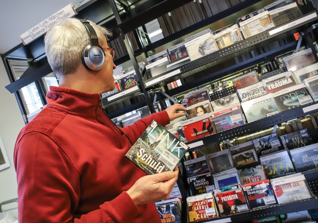 Ein Mann steht in der Stadtbücherei vor einem Regal mit Hörbüchern und trägt Kopfhörer. Er hält eine CD in der Hand.