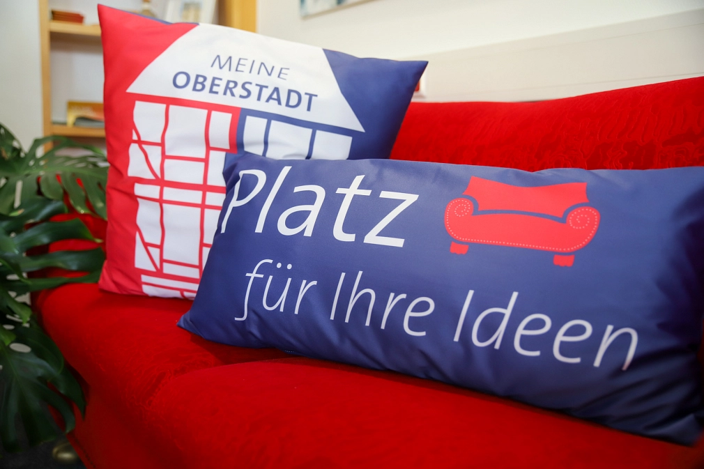 Auf einem roten Sofa sind zwei Kissen platziert: Das hintere trägt einen rot-blau-weißen Bezug mit einem stilisierten Fachwerkhaus. Im Dach stehen die Worte "Meine Oberstadt". Das vordere Kissen ist blau. Darauf gedruckt sind in weißer Schrift die Worte "Platz für deine Ideen" sowie ein rotes Sofa.