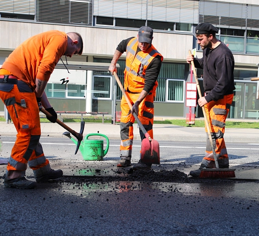 Drei Männer in orangefarbener Kleidung füllen mit Hacke und Schippe ein Schlagloch in der Straße auf.