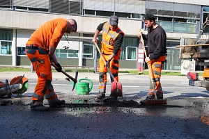 DBM flickt Schlaglöcher Drei Männer in orangefarbener Kleidung füllen mit Hacke und Schippe ein Schlagloch in der Straße auf.