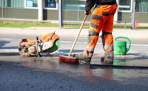 Ein Mann in orangefarbener Hose hat einen Besen in der Hand. Auf der Straße ist ein Schlagloch sowie weiteres Werkzeug und Geräte zu sehen.