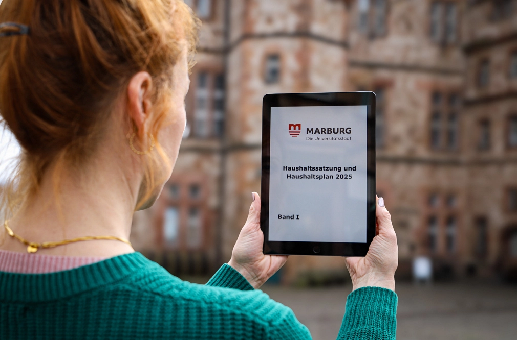 Eine Frau hält ein Tablet auf dem Marktplatz vor dem Marburger Rathaus in den Händen. Auf dem Display steht "Haushaltssatzung und Haushaltsplan 2025".&nbsp;&copy;&nbsp;Dennis Siepmann, Stadt Marburg
