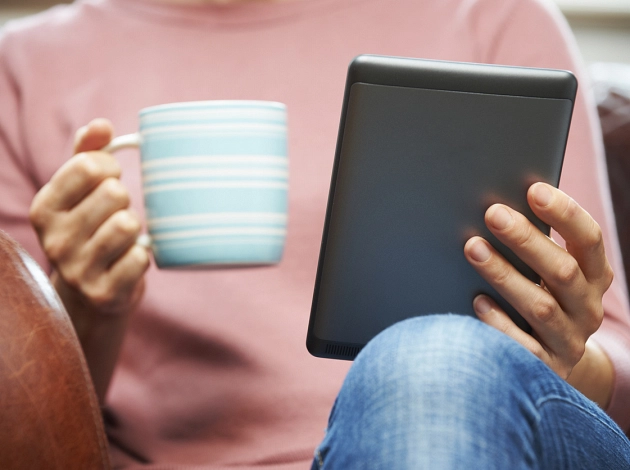 Eine Person sitzt in einem Sessel, in der einen Hand einen E-Book-Reader, in der anderen Hand eine Tasse