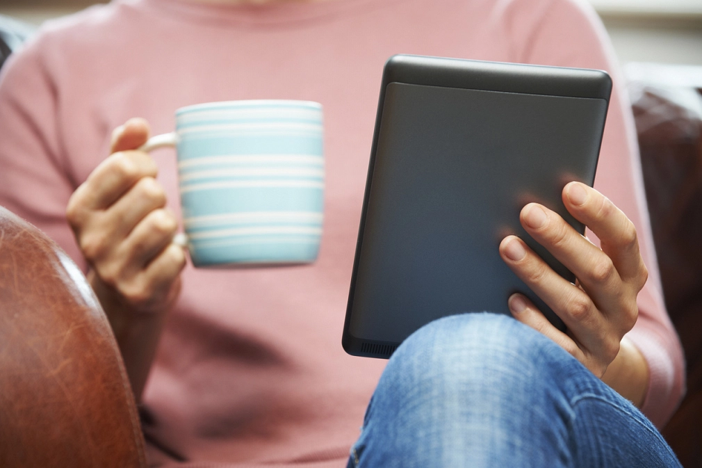 Digitale Stadtbücherei Eine Person sitzt in einem Sessel, in der einen Hand einen E-Book-Reader, in der anderen Hand eine Tasse © iStock/MachineHeadz