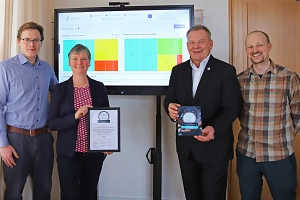 eGovSAD_Digital-Award 2024 Vier Personen stehen vor einem Bildschirm. Der Bildschirm zeigt eine Oberfläche mit mehreren Grafiken. Die zwei Personen in der Mitte halten eine Urkunde und einen Preis in der Hand. Es geht um den Digital-Award 2024.
