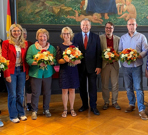 Ehrung Dienstjubiläen 2025 Einige Personen stehen in einem Raum, halbrund aufgestellt. Viele von Ihnen halten einen Blumenstrauß in der Hand.