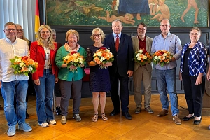 Ehrung Dienstjubiläen 2025 Einige Personen stehen in einem Raum, halbrund aufgestellt. Viele von Ihnen halten einen Blumenstrauß in der Hand.