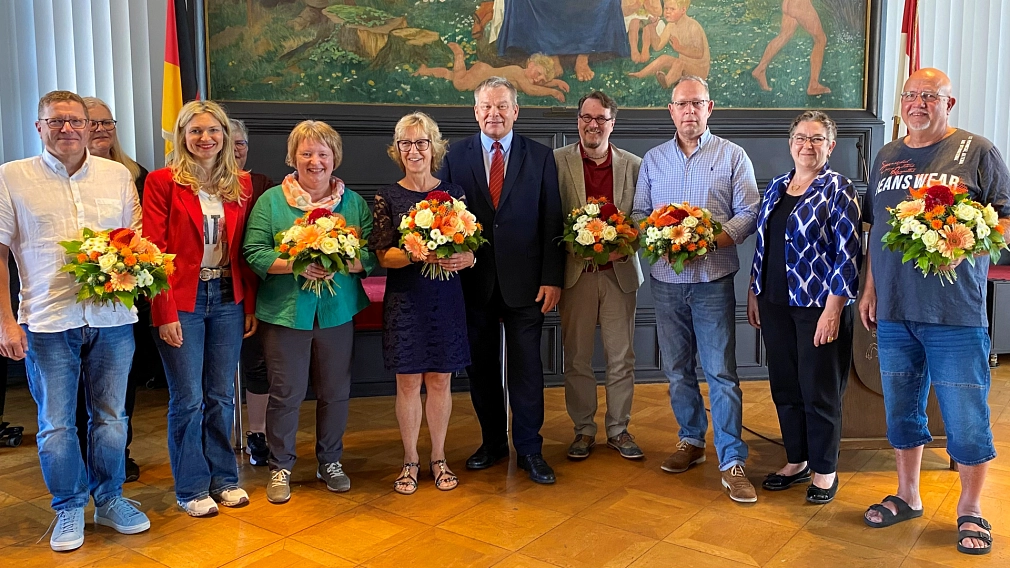Einige Personen stehen in einem Raum, halbrund aufgestellt. Viele von Ihnen halten einen Blumenstrauß in der Hand.