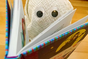 Ein selbstgemachtes Gespenst aus Mulltuch und mit Wackelaugen sitzt hinter einem aufgeschlagenen Buch und es wirkt, als würde es lesen.