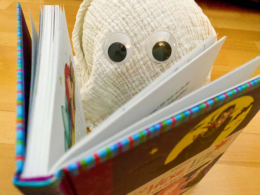 Ein Gespenst liest ein Buch in der Stadtbücherei Ein selbstgemachtes Gespenst aus Mulltuch und mit Wackelaugen sitzt hinter einem aufgeschlagenen Buch und es wirkt, als würde es lesen. © Simone Batz, Stadt Marburg