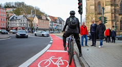 Das Bild zeigt einen Radfahrer an einer roten Fahrradampel. Im Hintergrund stehen Menschen und es fließt Autoverkehr.