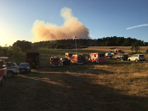 Am Himmel sind große Rauchwolken zu sehen. Im Vordergrund stehen viele Fahrzeuge der Feuerwehr auf einem Feld.