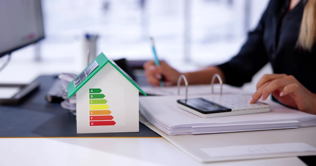 Ein Hausmodell auf einem Tisch. Auf dem Haus verschiedene Energieeffizienzklassen mit Farben. Eine Person am Tisch mit Taschenrechner.&nbsp;&copy;&nbsp;iStock/AndreyPopov