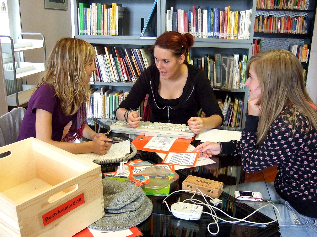 Escape Game in der Stadtbücherei Drei Mädchen sitzen zusammen an einem Tisch vor einem Bücherregal. Auf dem Tisch liegen verschiedene Gegenständen, unter anderem eine PC-Maus, ein Hut, beschriftete, orangefarbene Karten. Die mittlere Jugendliche spricht zu ihrer rechten Sitznachbarin, das dritte Mädchen hört lächelnd zu. © Universitätsstadt Marburg