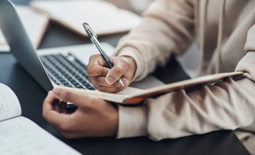 Fachdienst 13, Presse- und Öffentlichkeitsarbeit.jpg Zwie Hände einer Person, die am Schreibtisch sitzt, sind zu sehen. Die Person notiert etwas in ein Notizbuch. Auf dem Tisch ist noch ein Laptop zu sehen. © IStock / PeopleImages