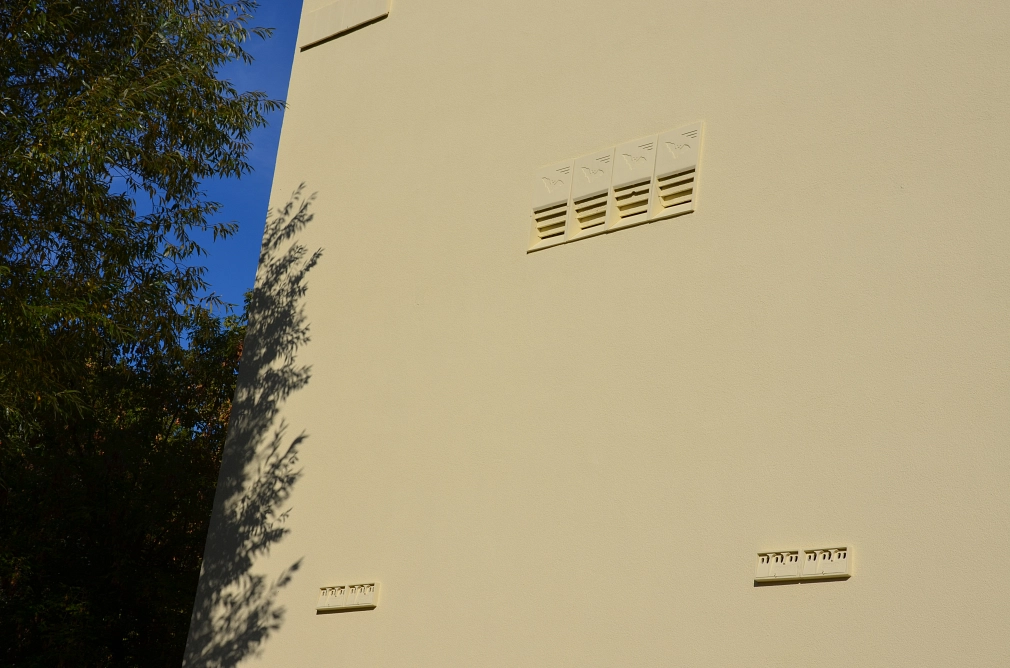 Mattgelbe Hausfassade, in die nebeneinander Fledermauskästen und in einigem Abstand darunter Sperlingsnisthilfen eingebaut wurden. Sie haben dieselbe Farbe wie die Fassade lockern den Fassadenanblick strukturell auf.