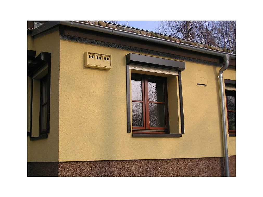 Mattgelbe Hausfassade mit einem Fledermauskasten und einer Sperlingsnisthilfe (beide in der ebenfalls mattgelb)