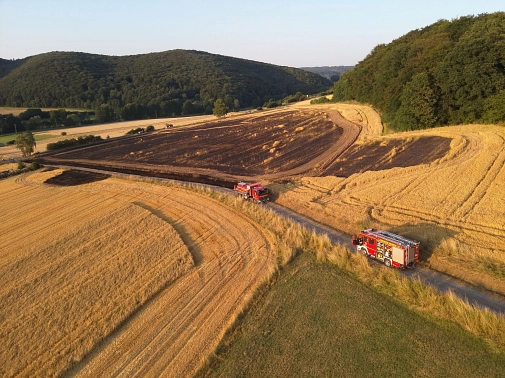 Das Bild zeigt ein Feld aus der Vogelperspektive, auf dem deutliche Brandspuren zu erkennen sind. Außerdem sieht man zwei Feuerwehrfahrzeuge.