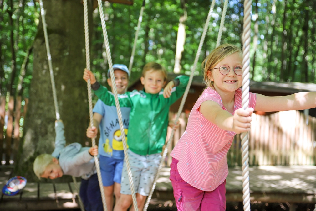 Ein Mädchen und drei Jungen halten sich an Tauen Seilen fest, sie balancieren auf hängenden Holzbalken im Stadtwald. Im Hintergrund sind Bäume zu sehen.