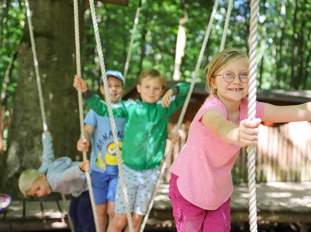 Ein Mädchen und drei Jungen halten sich an Tauen Seilen fest, sie balancieren auf hängenden Holzbalken im Stadtwald. Im Hintergrund sind Bäume zu sehen.