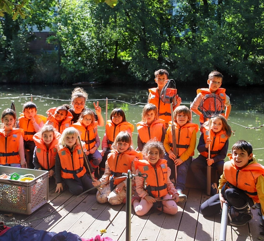 Floßprojekt 2025 Kinder stehen mit orangen Schwimmwesten auf einem Holz-Floß und schauen in die Kamera.