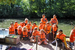 Floßprojekt 2025 Kinder stehen mit orangen Schwimmwesten auf einem Holz-Floß und schauen in die Kamera.