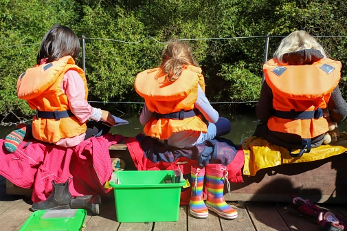 Drei Kinder mit Schwimmweste sitzen auf einer Bank auf dem Floß und schauen sich Bücher an.